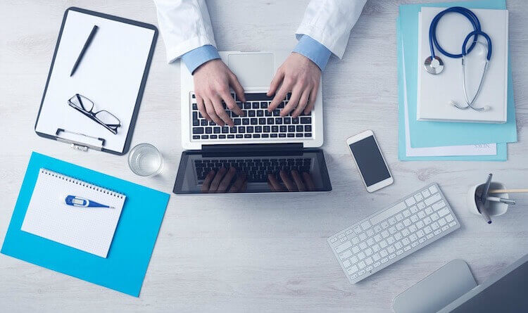 Médecin au travail sur un ordinateur avec stéthoscope, dossier patient et bloc note autour.